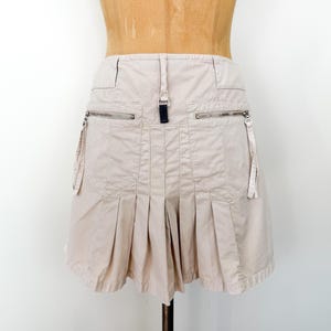 May include: A beige mini skirt with a pleated design and multiple pockets. The skirt features two zippered pockets on the front and a black tab detail at the waistband. The skirt is displayed on a mannequin.