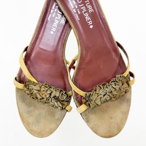 May include: A pair of open-toe sandals with a tan suede footbed and a brown leather insole. The sandals feature a ruffled accent across the top and thin yellow straps. The text "J PLINER" is visible on the insole.