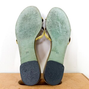 May include: A pair of yellow leather sandals, size 8M, with a thong-style design. The soles are green with wear marks, and the heels are black. The sandals are displayed on a wooden surface against a white background.