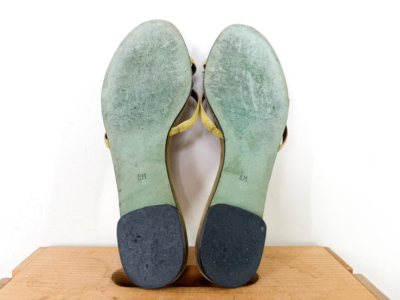 May include: A pair of yellow leather sandals, size 8M, with a thong-style design. The soles are green with wear marks, and the heels are black. The sandals are displayed on a wooden surface against a white background.