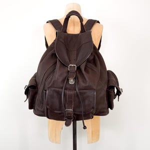 May include: A dark brown leather backpack featuring a large main compartment with a flap closure, a drawstring, and a metal buckle. The bag includes two side pockets with flap closures and a front pocket with a buckle, all secured with straps.