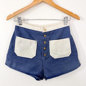 May include: High-waisted blue corduroy shorts featuring a contrasting cream-colored waistband and two large cream-colored patch pockets on the front. The shorts are secured with a vertical row of five small, round, light-colored wooden buttons.
