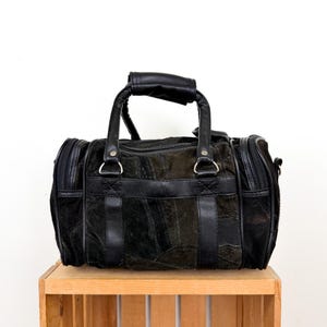 May include: Black duffel bag with a patchwork design. The bag has a top handle and two side zippers. The bag is made of suede and leather. The bag is sitting on a wooden crate.