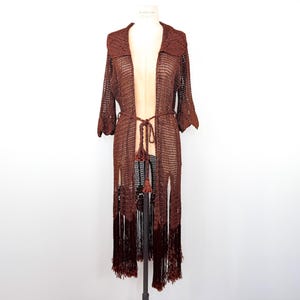 May include: A long, open-front brown crochet cardigan with a collared neckline and three-quarter sleeves. The cardigan features a matching belt and fringe detailing along the bottom. The garment is displayed on a mannequin.