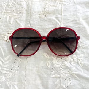 May include: Red-framed sunglasses with dark lenses. The sunglasses have a round shape and a thick frame. The lenses are gradient, transitioning from dark at the top to lighter at the bottom. The sunglasses are set against a white, embroidered fabric.