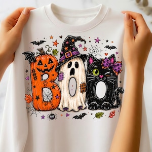 May include: A white crewneck sweatshirt featuring a colorful Halloween graphic. The design displays the word BOO, where the B is an orange jack-o'-lantern, the O is a white ghost wearing a witch hat, and the second O is a black cat with a purple bow.