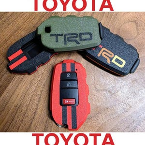 May include: A collection of Toyota TRD key fobs in red, green, and black. The red fob has black stripes and buttons for lock, unlock, and hold. The green fob has the letters TRD. The word TOYOTA is displayed in red.