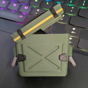 May include: An olive green case with a hinged lid, featuring a yellow stripe and black straps. The case has a cross design on the front and grey side latches. The case is on a laptop keyboard.