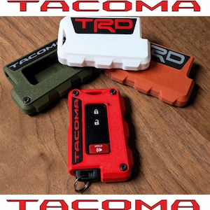May include: Four rectangular key fob covers for a Toyota Tacoma, each featuring a textured finish and the TRD or Tacoma logo. The covers are available in white, red, olive green, and orange, with one red cover installed on a black key fob.