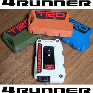 Puede incluir: Un llavero blanco con botones negros y detalles rojos, incluyendo la palabra "HOLD". Tres cubiertas de llavero de colores en naranja, azul y verde, cada una con "TRD" en rojo. El texto "4RUNNER" está arriba y abajo de los artículos.