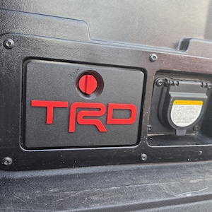Op de afbeelding: Een zwart rechthoekig opbergvakdeurtje met het rode TRD-logo en een rood rond draaimechanisme met het opschrift OPEN. Naast het vak bevindt zich een zwart stopcontact met een beschermkap en een geel waarschuwingslabel.