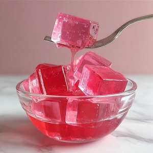 Può includere: Una ciotola di vetro trasparente piena di dessert di gelatina rossi a forma di cubo. Una forchetta tiene un cubo, con un rivolo di gelatina che gocciola. Lo sfondo è rosa tenue e la ciotola è su una superficie di marmo bianco.