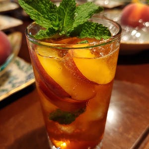 May include: A refreshing cocktail in a clear glass, featuring peach slices and fresh mint sprigs. The drink has a golden hue, with visible bubbles, and is garnished with a vibrant green mint leaf. The glass sits on a wooden surface.