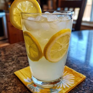 May include: A clear glass filled with a pale yellow lemonade, ice cubes, and three bright yellow lemon slices. One lemon slice is perched on the rim, while two others are submerged inside the glass. The drink rests on a yellow floral napkin.