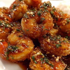 May include: A plate of fried shrimp, glistening with a rich, brown sauce. The shrimp are sprinkled with chopped green herbs and nuts. The dish is presented on a white plate, highlighting the golden-brown hue of the seafood.