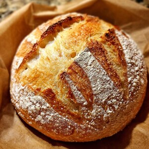 May include: A round loaf of freshly baked bread, resting on brown parchment paper. The crust is golden brown with diagonal scoring and a dusting of white flour. The bread appears rustic and homemade, perfect for a bakery or kitchen.