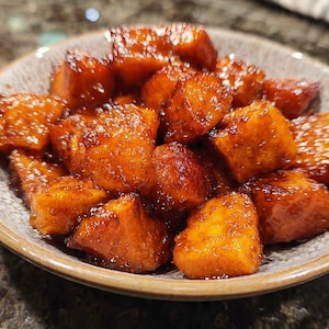 Puede incluir: Primer plano de un cuenco lleno de boniatos glaseados y caramelizados. Los boniatos están cortados en cubos y tienen un color ámbar brillante. El cuenco es gris con un borde texturizado.