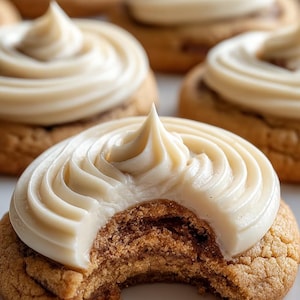 Puede incluir: Primer plano de varias galletas glaseadas. Las galletas tienen un exterior marrón claro y texturizado y un glaseado blanco cremoso arremolinado en la parte superior. Una galleta tiene un mordisco, revelando el interior marrón suave. Las galletas están dispuestas sobre una superficie blanca.