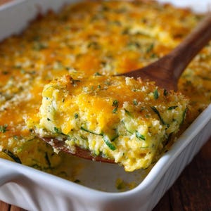 Puede incluir: Una fuente rectangular blanca llena de calabacín y queso horneados. Una cuchara de madera levanta una porción de la cazuela, mostrando las capas de calabacín, queso y cobertura de pan rallado. El plato está sobre una superficie de madera.
