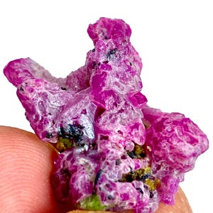 May include: A raw, vibrant magenta-colored mineral specimen with a crystalline, textured surface. The cluster features irregular, jagged formations with small, dark inclusions and patches of yellowish-green mineral deposits embedded within the structure.