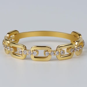 Könnte beinhalten: Ein goldfarbener Ring mit einem Kettenglied-Design. Der Ring hat rechteckige Glieder, die jeweils mit kleinen, klaren Edelsteinen besetzt sind. Der Ring ist vor einem schlichten, hellen Hintergrund platziert, der seine polierte Oberfläche und die Details hervorhebt.