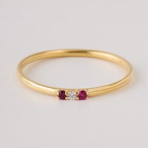 May include: A slender gold ring with a small diamond and two red gemstones. The ring's band is thin, with a minimalist design. The gemstones are set in a row, adding a touch of colour and sparkle. A simple and elegant piece of jewellery.