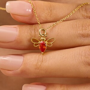 May include: A gold-toned necklace featuring a small bee-shaped pendant. The bee has delicate wire-frame wings and a single, vibrant, pear-shaped orange-red gemstone set in the center of its body, suspended from a thin, matching gold-toned chain.
