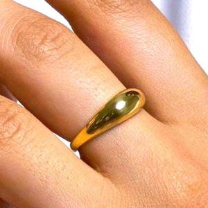 Könnte beinhalten: Ein polierter goldfarbener Ring mit einer glatten, sich verjüngenden Kuppelform. Das Band besteht aus einer massiven, durchgehenden Metallkonstruktion mit einer hochglänzenden Oberfläche, die sich sanft um den Finger wölbt und schlicht wirkt.