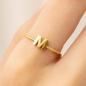 May include: A gold-colored ring with the letter "M" on the finger of a person. The ring has a twisted band design. The ring is a piece of jewelry and is likely made of metal.