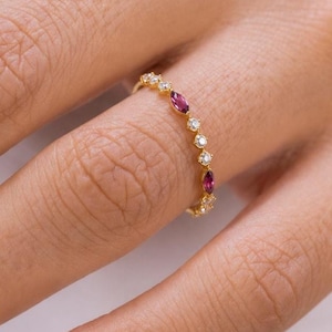 Könnte beinhalten: Ein goldener Ring mit einer feinen Anordnung aus abwechselnd im Marquise-Schliff geschliffenen magentafarbenen Edelsteinen und kleinen runden, klaren Steinen. Die dünne Metallschiene hält die Steine in einer Reihe und bildet ein Schmuckstück.