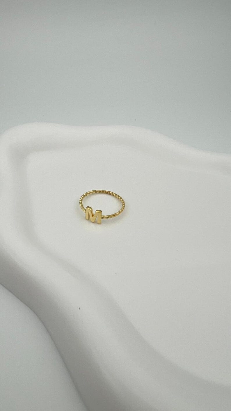 May include: A gold-colored ring with the letter "M" in the center. The ring has a twisted band design. The ring is displayed on a white, curved surface.