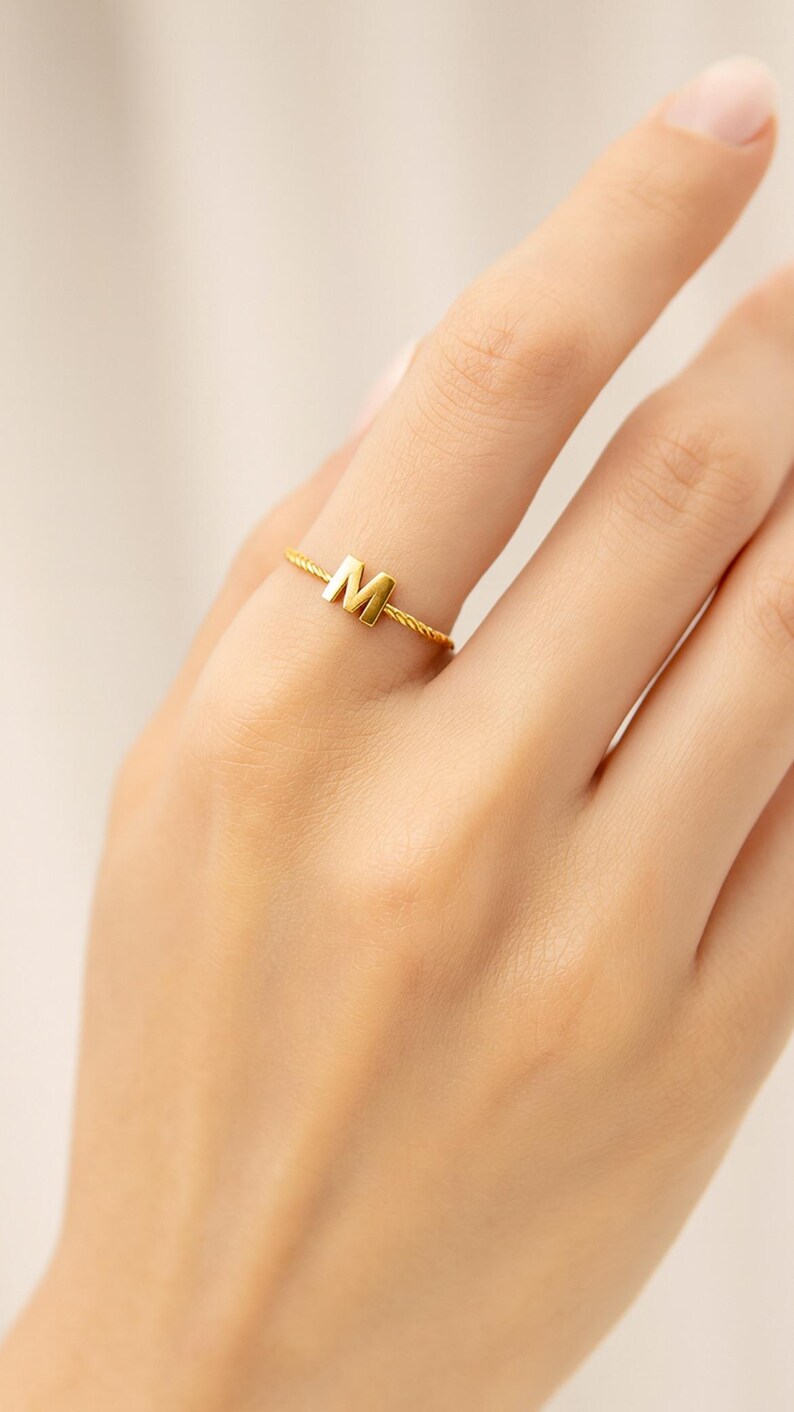 May include: A gold-colored ring with the letter "M" on the finger of a person. The ring has a twisted band design. The ring is a piece of jewelry and is likely made of metal.