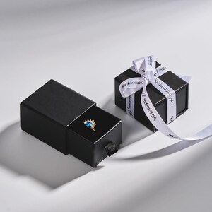 May include: A black jewelry box with a ring inside, next to a wrapped gift box. The ring features a gold band and a blue gemstone. The gift box is tied with a white ribbon with black text.