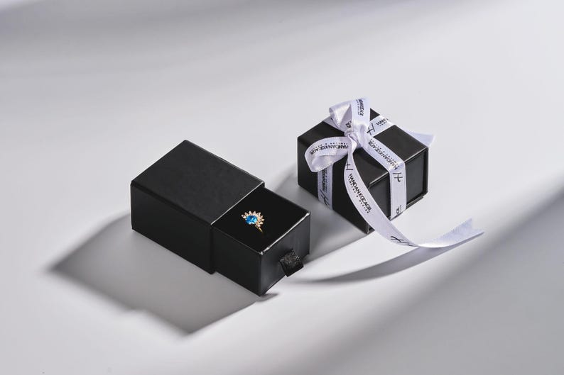 May include: A black jewelry box with a ring inside, next to a wrapped gift box. The ring features a gold band and a blue gemstone. The gift box is tied with a white ribbon with black text.