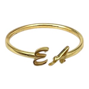 May include: A gold-colored ring with the letters "E" and "A" in a script font. The ring is open at the front, with the letters forming the focal point. The ring is likely made of gold or a gold-colored metal.