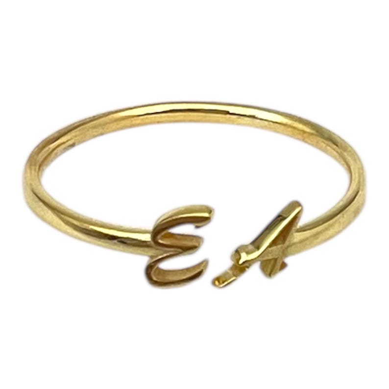 May include: A gold-colored ring with the letters "E" and "A" in a script font. The ring is open at the front, with the letters forming the focal point. The ring is likely made of gold or a gold-colored metal.