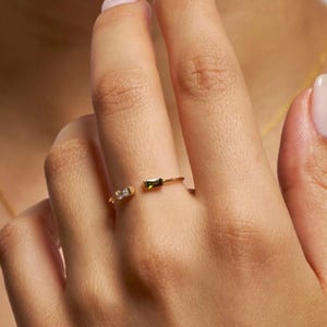 May include: A gold open-style ring featuring two baguette-cut stones. One stone is clear and colourless, while the other is a deep olive green. The thin, polished gold band remains open at the centre, showcasing the two distinct rectangular gemstones.