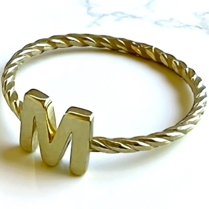 May include: A gold-colored ring with a twisted band and a raised letter "M". The ring is made of a shiny, metallic material and is set against a white background. The ring's design is simple yet elegant, suitable for everyday wear.