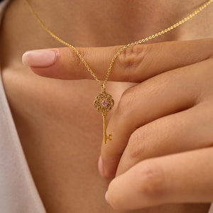 May include: A gold-colored key pendant necklace. The key has an ornate design with a pink gemstone in the center. The necklace is held by a person's hand, showcasing the delicate chain and pendant. The key pendant is a symbol of unlocking possibilities.