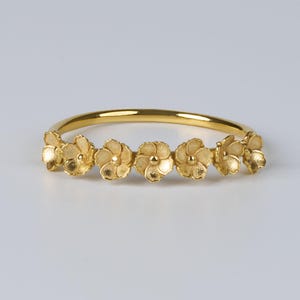Könnte beinhalten: Ein goldfarbenes Armband mit sechs detaillierten Blumenmotiven. Die Blumen sind gleichmäßig um das Band angeordnet, jede mit einem kleinen, erhabenen Zentrum. Das Armband hat eine glatte, polierte Oberfläche, und die Blumen haben ein strukturiertes Aussehen.