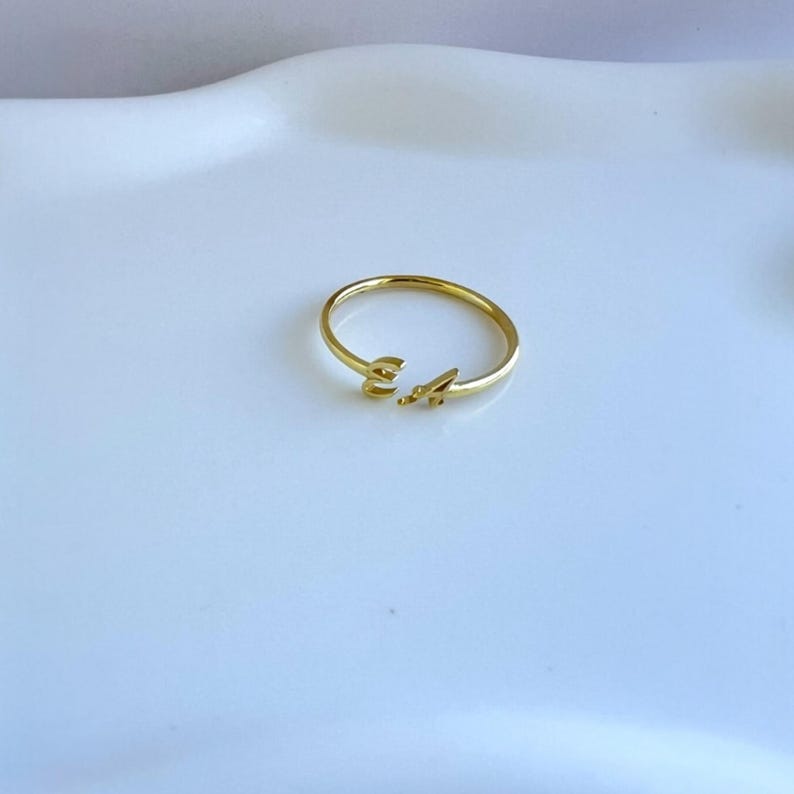 May include: A delicate gold ring featuring the letters "E" and "A". The ring has a minimalist design with the letters placed on the band. The ring is set against a white background.