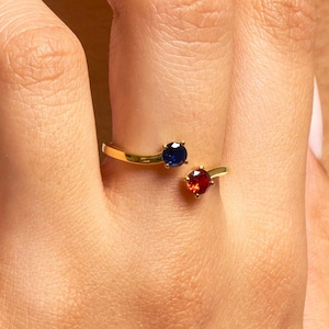 Könnte beinhalten: Goldfarbener Ring mit Bypass-Design, besetzt mit einem runden, dunkelblauen Edelstein und einem runden, roten Edelstein. Der Ring wird an einem Finger gezeigt.