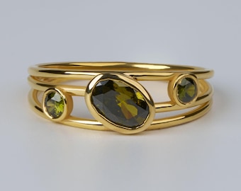 May include: A gold ring featuring three bands connected by three olive green gemstones. The center stone is a large oval shape, while the two smaller stones on either side are round. The metal has a polished finish and a warm yellow tone.