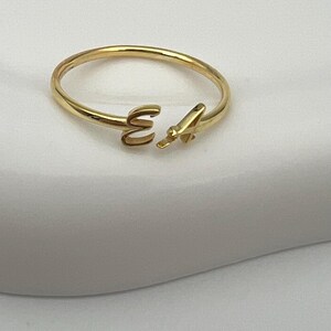 May include: A gold-colored open ring with the letters "E" and "A" in a delicate, minimalist design. The ring is made of a shiny, smooth metal and is set against a soft, white background. A simple and elegant piece of jewelry.