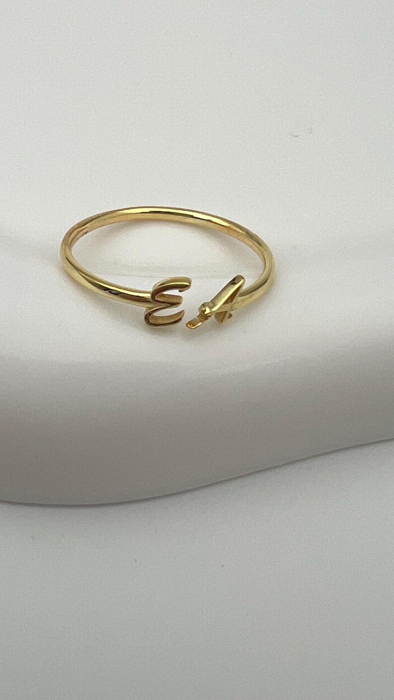 May include: A gold-colored open ring with the letters "E" and "A" in a delicate, minimalist design. The ring is made of a shiny, smooth metal and is set against a soft, white background. A simple and elegant piece of jewelry.