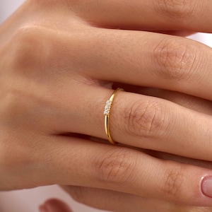 Può includere: Un sottile anello a fascia in oro con tre piccole pietre chiare a taglio rotondo disposte in fila orizzontale. Il metallo ha una finitura lucida e riflettente, e le pietre sono posizionate centralmente sulla parte superiore del sottile cerchio.