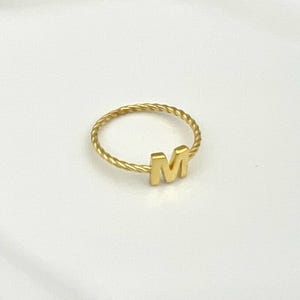 May include: A gold-colored ring with a twisted band and the letter "M" in a block font. The ring is set against a white background, highlighting its simple design and metallic sheen.