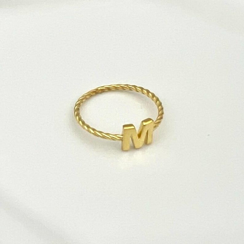May include: A gold-colored ring with a twisted band and the letter "M" in a block font. The ring is set against a white background, highlighting its simple design and metallic sheen.