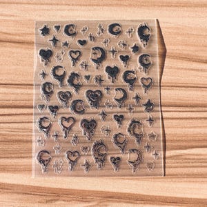 May include: A clear stamp sheet featuring black designs of crescent moons, hearts, stars, and crosses. The designs have a dripping effect, creating a unique aesthetic. The sheet is on a wooden surface.