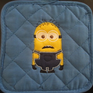 May include: A blue quilted pot holder with an embroidered yellow minion wearing blue overalls. The minion has a black mustache and goggles.